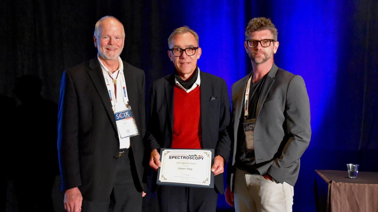 Three men on stage under blue light; center man in red sweater holds certificate