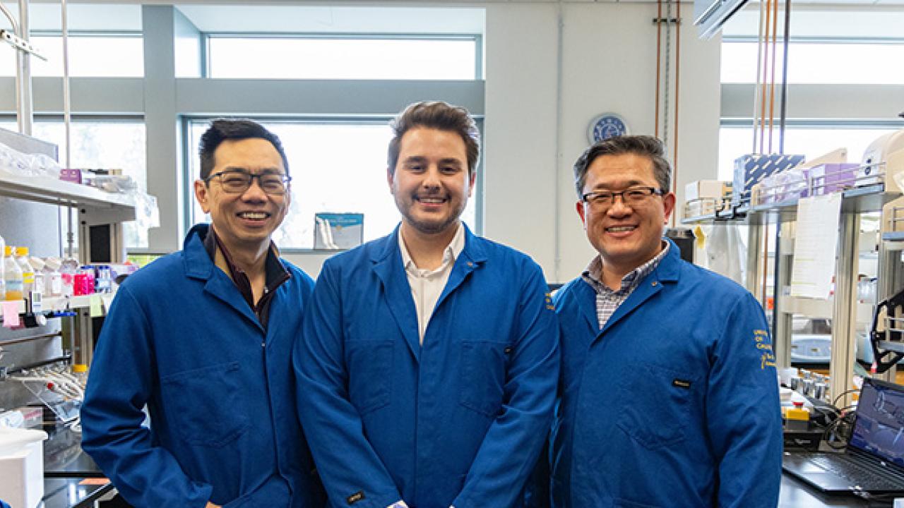 Three smiling researchers in blue lab coats standing in a bright laboratory.